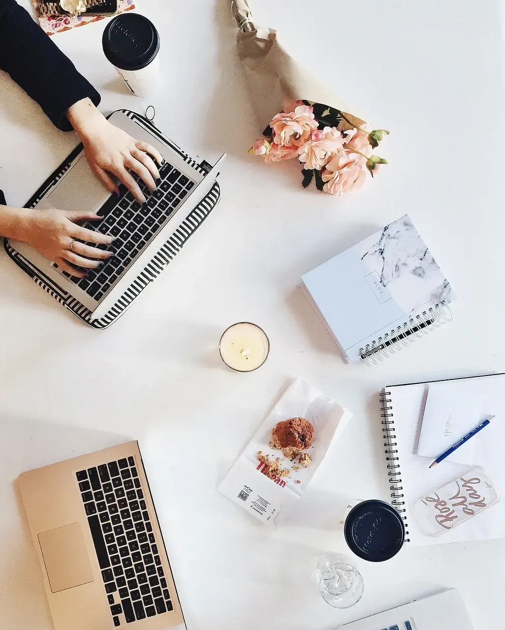 desk with person typing on laptop, two notebooks, another laptop, a candle, and an eaten muffin